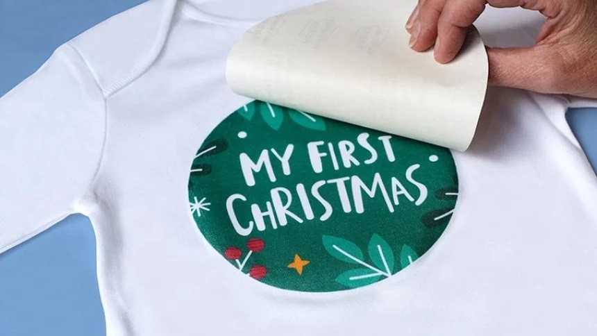 Main en train de réaliser un transfert textile sur t-shirt blanc avec un design de Noël