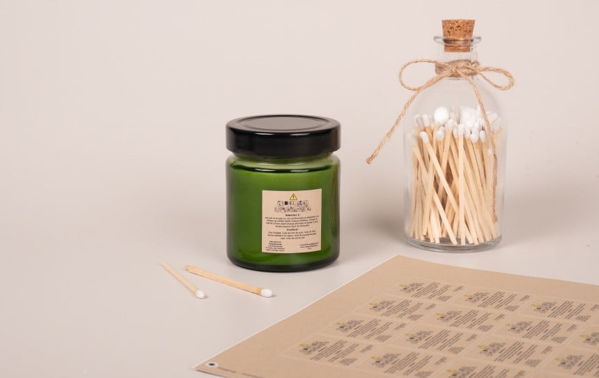 Etiquette règlementaire pour bougie en papier kraft collée sur un contenant vert pour bougie avec des planches d'étiquettes à côté