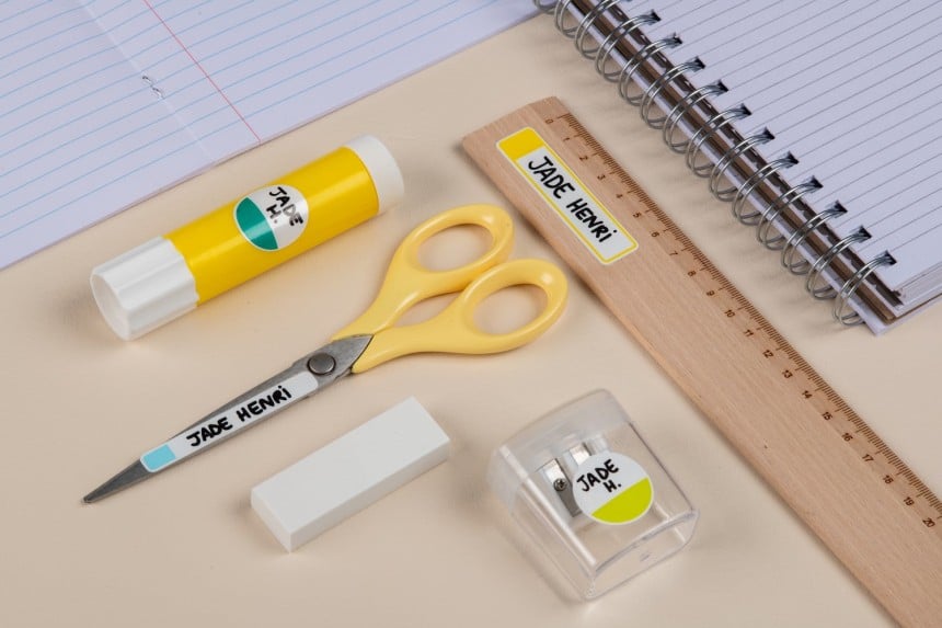 Etiquettes plastifiées collées sur une règle, un tube de colle, des ciseaux et un taille-crayon.