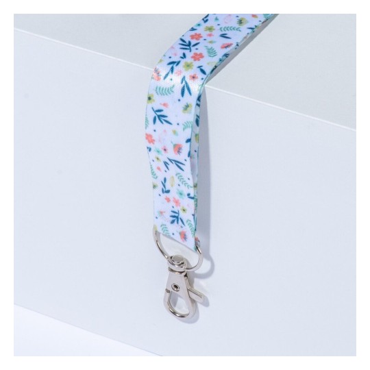 L'image montre un porte-clés ou un cordon décoré de motifs floraux colorés, attaché à un crochet en métal, reposant sur une surface blanche.