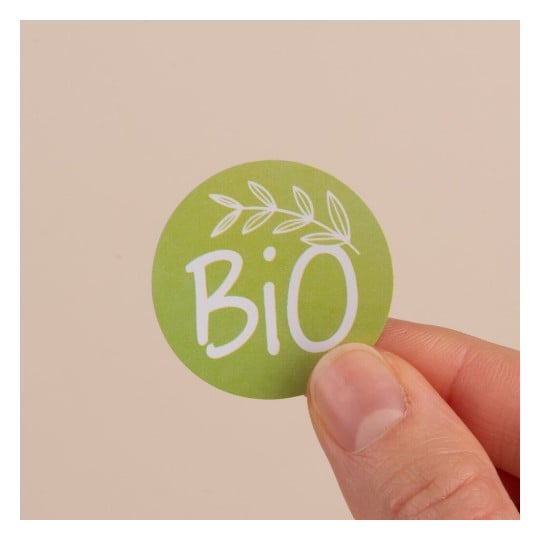 L'image montre un autocollant circulaire vert avec le mot 'BiO' en blanc, entouré de feuilles, tenue par une main sur un fond beige.