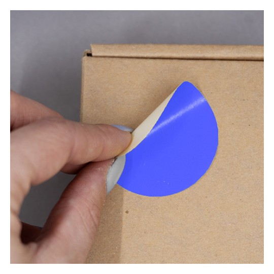 L'image montre une main qui décolle un autocollant bleu d'une boîte en carton. Le sticker a une partie de couleur beige qui est encore collée.