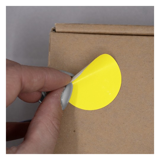 L'image montre une main qui décolle un autocollant jaune rond d'une boîte en carton.