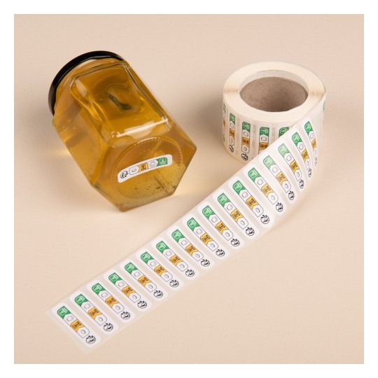 L'image montre un pot de miel en verre avec un couvercle noir, à côté d'un rouleau d'étiquettes autocollantes. Les étiquettes présentent des icônes en vert et jaune, probablement pour des informations de sécurité ou d'étiquetage.