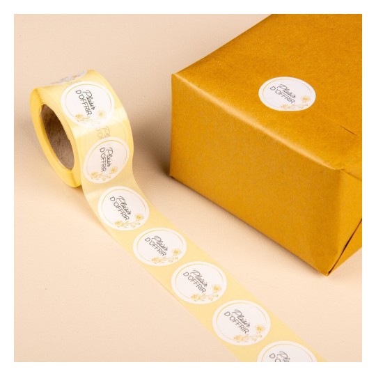 L'image montre un rouleau de scotch avec des étiquettes blanches et une boîte cadeau dorée sur une surface beige.