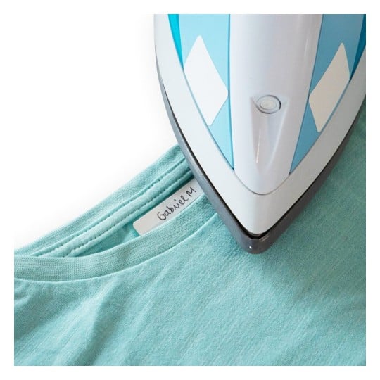 L'image montre un fer à repasser bleu placé sur un t-shirt turquoise. Le t-shirt a une étiquette avec le nom Gabriel M.