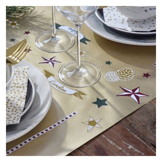 L'image présente une nappe dorée décorée avec des étoiles, des cônes de pin et des étiquettes personnalisées comme 'Caroline'. Il y a deux verres à vin et de la vaisselle élégante, incluant des assiettes et des serviettes à motifs dorés.