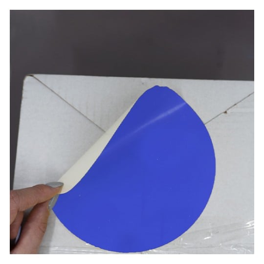 L'image montre une personne tenant un cercle bleu qui semble être une étiquette ou un autocollant, en train d'être décollé d'un carton blanc.