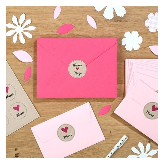 L'image montre plusieurs enveloppes colorées, y compris une enveloppe rose avec un symbole sur le devant, ainsi que des fleurs en papier et des feuilles découpées sur une surface en bois.