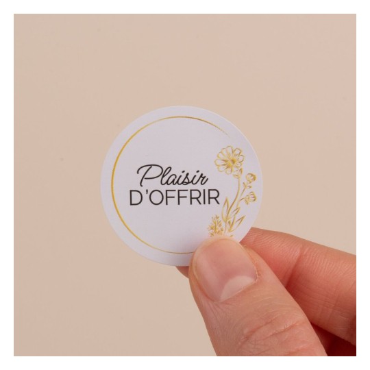 Il s'agit d'une étiquette ronde tenant dans une main, avec le texte 'Plaisir d'Offrir' et un motif floral doré sur un fond clair.