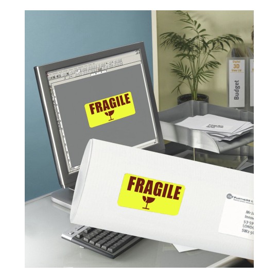 L'image montre un ordinateur de bureau avec un écran affichant un étiquette 'FRAGILE' et un tube ou une boîte avec la même étiquette au premier plan. Le bureau est organisé avec des documents et une plante en arrière-plan.