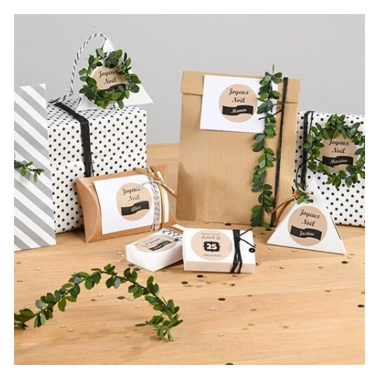 L'image montre divers cadeaux joliment emballés avec des étiquettes portant des noms. Les paquets sont décorés de verdure et sont disposés sur une table en bois.