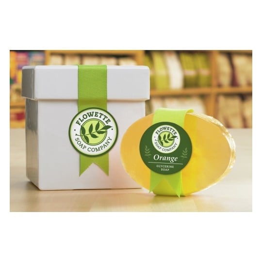 L'image montre une boîte blanche avec un ruban vert portant le logo de 'Flowette Soap Company' et un savon en glycérine de couleur orange à côté.