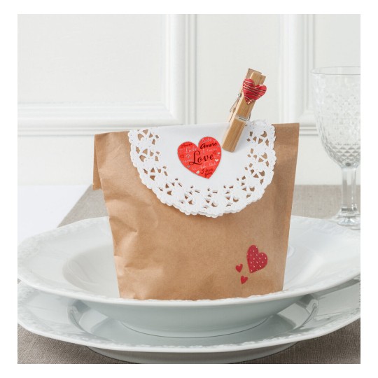 L'image montre un sac en papier décoré avec une dentelle blanche, un cœur rouge et une pince à linge. Il est posé sur une assiette, suggérant un thème romantique ou de célébration.