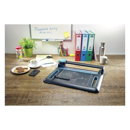 L'image montre un bureau avec une machine de découpe, des fournitures de bureau, une tasse de café, des biscuits, un ordinateur portable et des classeurs colorés. Un tableau blanc indique un devoir de cour de récréation.