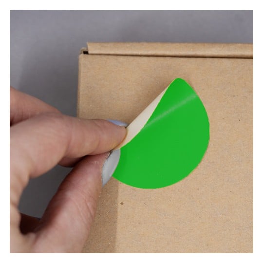 L'image montre une personne en train de décoller un autocollant vert d'un emballage en carton. Le décor est minimaliste, mettant en valeur le geste de la main et le sticker.