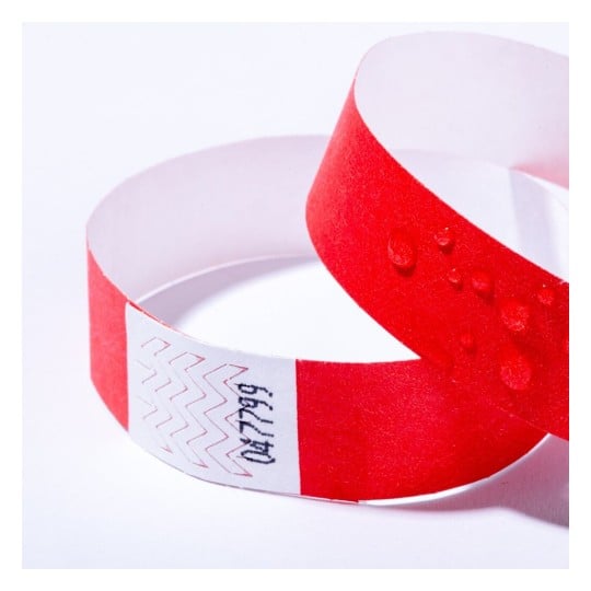 L'image montre un bracelet en papier rouge et blanc, avec des gouttes d'eau sur sa surface. Il semble utilisé pour l'identification ou l'accès à un événement.