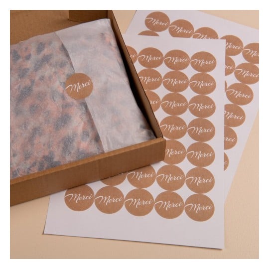 L'image montre une boîte en carton contenant un papier kraft avec une étiquette 'Merci', ainsi que des feuilles de papier décorées de cercles portant le même texte.