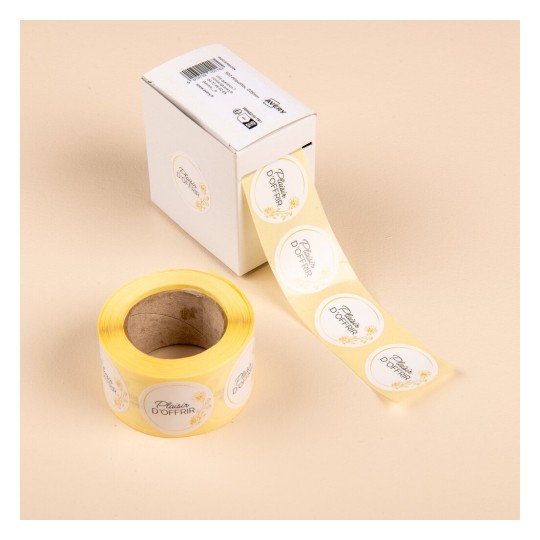 L'image montre une boîte blanche contenant des rouleaux d'étiquettes autocollantes jaunes avec le texte 'Plaisir d'Offrir'. Les étiquettes sont déroulées et enroulées sur un rouleau. L'arrière-plan est de couleur beige.