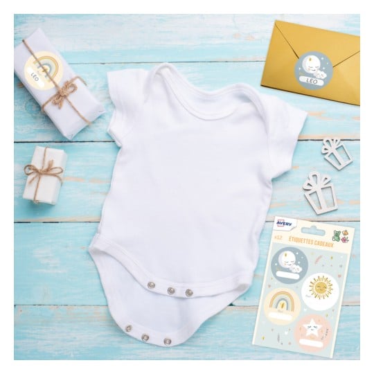 L'image montre un body blanc pour bébé placé sur une surface en bois teintée de bleu, entouré de petits cadeaux emballés et d'une enveloppe dorée avec des étiquettes diverses.