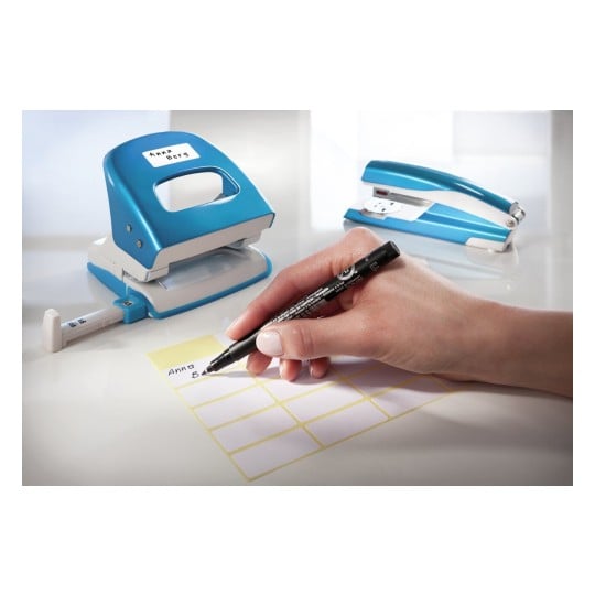 L'image montre un bureau avec une perforatrice bleue, une agrafeuse et une main utilisant un marqueur noir pour écrire sur une étiquette jaune.