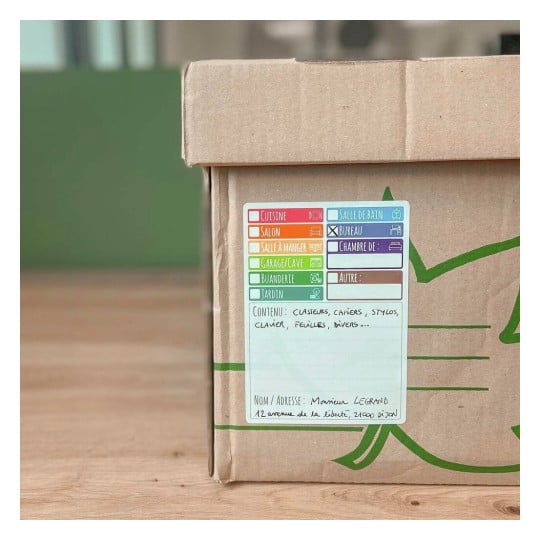Une boîte en carton avec une étiquette indiquant son contenu et l'adresse de livraison. Les catégories sur l'étiquette incluent cuisine, salon, salle à manger, garage, buanderie, jardin, et d'autres, avec des éléments comme des classeurs et des stylos listés comme contenu.