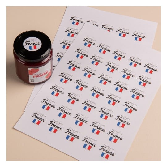L'image montre un pot de confiture de fraise à côté de plusieurs feuilles avec des étiquettes imprimées qui disent 'Fabrique en France' en plusieurs exemplaires.
