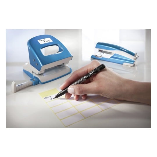 L'image montre une main utilisant un stylo noir pour écrire sur une étiquette jaune, à côté d'une perforatrice et d'une agrafeuse bleue sur une surface blanche.