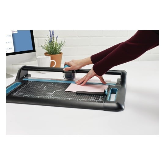 L'image montre une personne utilisant un outil de découpe sur un bureau, avec un écran d'ordinateur et une tasse à proximité.