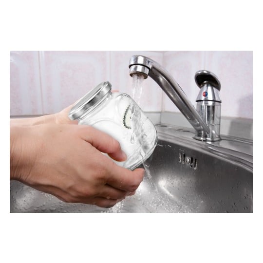 Une personne lave un bocal en verre sous un robinet dans un évier. L'eau coule du robinet sur le bocal.