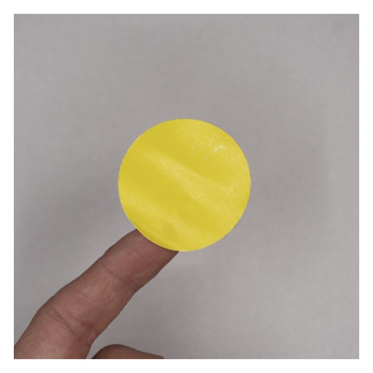 Il s'agit d'un cercle jaune sur un doigt, sur un fond gris. Le cercle semble en matériau lisse avec une texture légère.