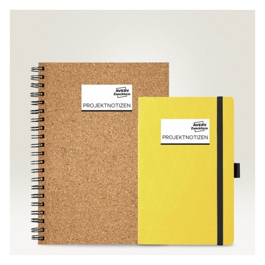 L'image montre deux carnets de notes. Le carnet de gauche est en liège et de format A5, tandis que celui de droite est jaune avec une couverture lisse et un élastique de maintien.