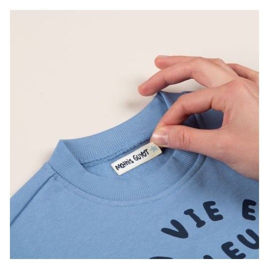 L'image montre un pull bleu avec une étiquette portant le nom 'Marin's Guyot' et un motif de texte noir qui dit 'VIE E'. Une main tient le col du pull.
