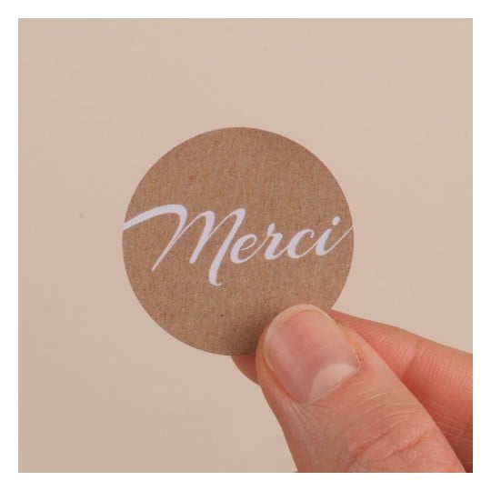 L'image montre un petit cercle en papier avec le mot 'Merci' écrit en blanc sur un fond brun. La main qui tient le cercle est visible.