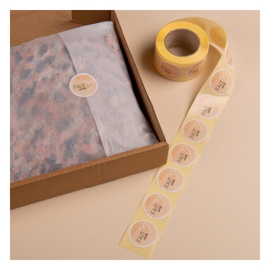 Cette image montre une boîte en carton avec du papier de soie à l'intérieur, étiquetée avec un autocollant rond. À côté, un rouleau d'étiquettes autocollantes est visible.