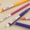 L'image montre plusieurs stylos colorés étiquetés avec le nom 'Laura S.' sur un fond en bois.