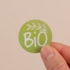 L'image montre un autocollant circulaire vert avec le mot 'BiO' en blanc, entouré de feuilles, tenue par une main sur un fond beige.