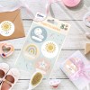 Cette image montre des étiquettes cadeaux colorées avec des illustrations joyeuses, un enveloppe en papier kraft, une petite boîte cadeau et d'autres accessoires sur une surface en bois.