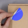 L'image montre une main qui décolle un autocollant bleu d'une boîte en carton. Le sticker a une partie de couleur beige qui est encore collée.