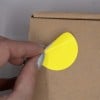 L'image montre une main qui décolle un autocollant jaune rond d'une boîte en carton.