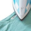L'image montre un fer à repasser bleu placé sur un t-shirt turquoise. Le t-shirt a une étiquette avec le nom Gabriel M.