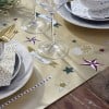 L'image présente une nappe dorée décorée avec des étoiles, des cônes de pin et des étiquettes personnalisées comme 'Caroline'. Il y a deux verres à vin et de la vaisselle élégante, incluant des assiettes et des serviettes à motifs dorés.