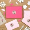 L'image montre plusieurs enveloppes colorées, y compris une enveloppe rose avec un symbole sur le devant, ainsi que des fleurs en papier et des feuilles découpées sur une surface en bois.