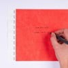 L'image montre une main écrivant sur un papier rouge avec des lignes et des étiquettes blanches sur le côté gauche. Les mots 'YOUR TEXT HERE' et 'VOTRE TEXTE ICI' sont visibles.