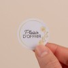 Il s'agit d'une étiquette ronde tenant dans une main, avec le texte 'Plaisir d'Offrir' et un motif floral doré sur un fond clair.