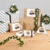 L'image montre divers cadeaux joliment emballés avec des étiquettes portant des noms. Les paquets sont décorés de verdure et sont disposés sur une table en bois.