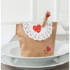 L'image montre un sac en papier décoré avec une dentelle blanche, un cœur rouge et une pince à linge. Il est posé sur une assiette, suggérant un thème romantique ou de célébration.