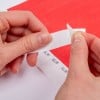 L'image montre une personne qui retire délicatement un morceau de papier adhésif d'une bande, avec un fond rouge.