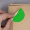 L'image montre une personne en train de décoller un autocollant vert d'un emballage en carton. Le décor est minimaliste, mettant en valeur le geste de la main et le sticker.