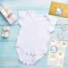 L'image montre un body blanc pour bébé placé sur une surface en bois teintée de bleu, entouré de petits cadeaux emballés et d'une enveloppe dorée avec des étiquettes diverses.